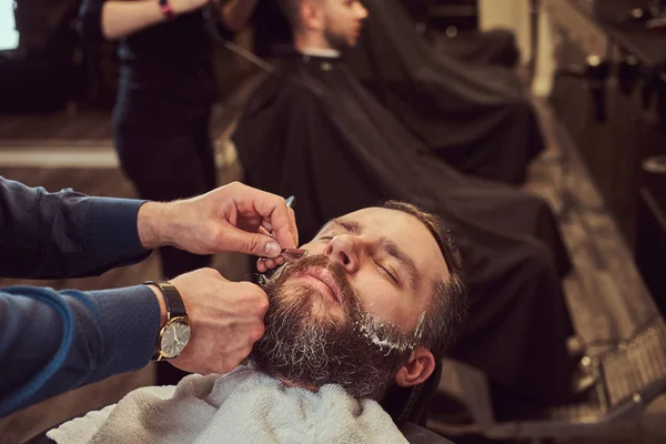 Homme barbu assis dans un fauteuil dans un salon de coiffure tandis que le coiffeur se rase la barbe avec un rasoir dangereux . — Photo