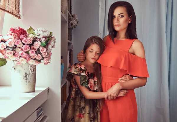 Fashion family. A tender mother and cute daughter at home. Little girl hugging her mother, standing near the window. — Stock Photo, Image