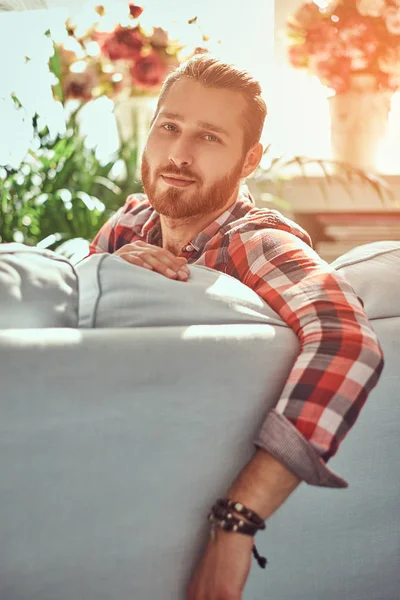 Молодой красивый рыжий мужчина-модель сидит на диване в комнате с интерьером лофта . — стоковое фото