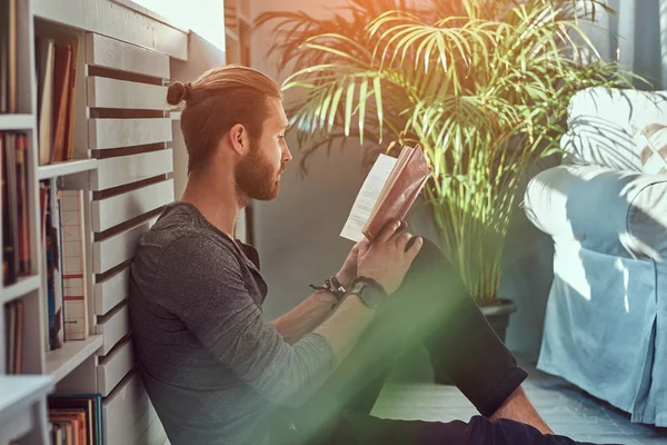 Retrato de un guapo estudiante pelirrojo con ropa casual, sentado en un piso en una casa mientras lee un libro, mirando a una cámara . — Foto de Stock
