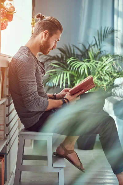 Bello studente rossa elegante in abiti casual, seduto su una sedia in una casa durante la lettura di un libro . — Foto Stock