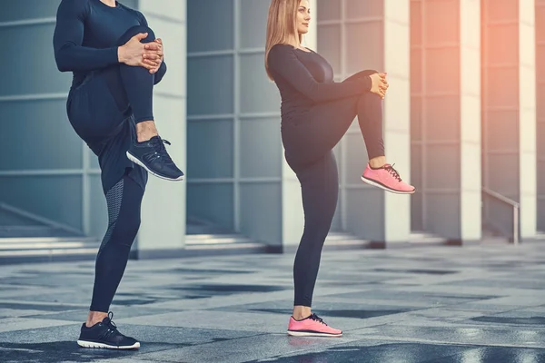 Joven pareja de fitness en ropa deportiva, haciendo estiramientos en la ciudad moderna contra un rascacielos. concepto de estilo de vida saludable . — Foto de Stock