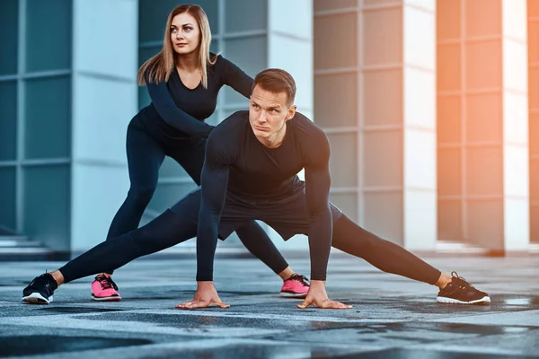 Joven pareja de fitness en ropa deportiva, haciendo estiramientos mientras se prepara para el ejercicio serio en la ciudad moderna contra un rascacielos . — Foto de Stock