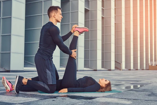 Young fitness couple in a sportswear, doing stretching while preparing for serious exercise in the modern city against a skyscraper. Healthy lifestyle concept. — Stock Photo, Image