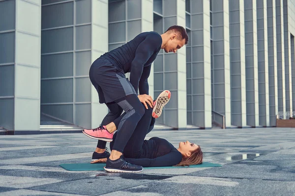 Joven pareja de fitness en ropa deportiva, haciendo estiramientos mientras se prepara para el ejercicio serio en la ciudad moderna contra un rascacielos. concepto de estilo de vida saludable . — Foto de Stock