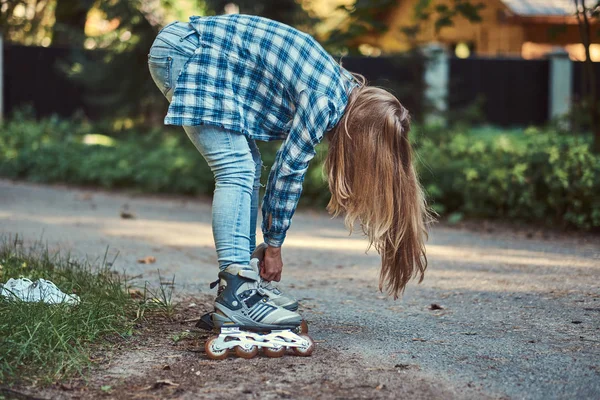 Красивая девушка с длинными волосами в флисовой рубашке и джинсах надевает ролики в парке в летний день . — стоковое фото