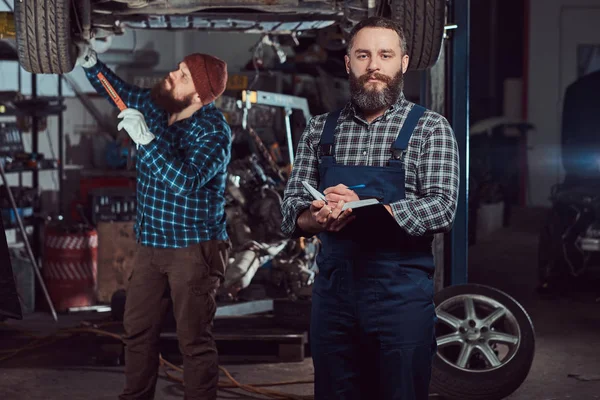 Two bearded brutal mechanics repair a car on a lift in the garage.