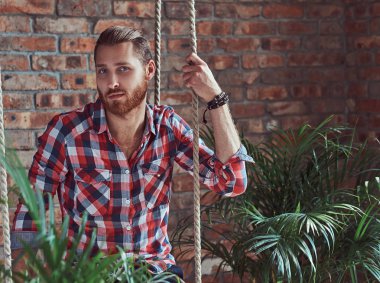 Genç yakışıklı Kızıl saçlı model adam bir çatı katı iç odadaki bir salıncak üzerinde oturur.