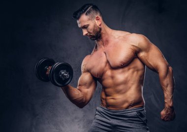 Uzun şık gömleksiz vücut geliştirmeci pazı ile bir stüdyoda ayakta bir dumbbell egzersiz yapıyor spor şort giymiş. Karanlık bir arka plan üzerinde izole.