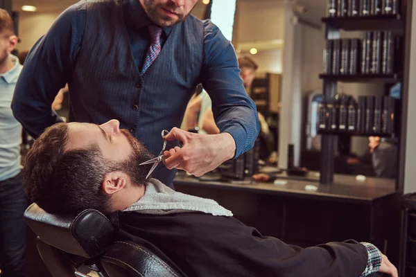 Профессиональный парикмахер моделирует бороду с ножницами и расческой в парикмахерской . — стоковое фото