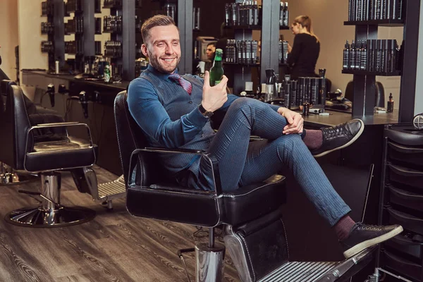 Ein professioneller Friseur sitzt auf einem Sessel und ruht sich nach der Arbeit mit einer Bierflasche in der Hand aus. — Stockfoto