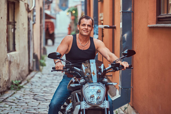 A handsome brutal biker dressed in a black t-shirt and jeans, with a charming smile, sitting on a motorcycle, in a narrow old Europe street.