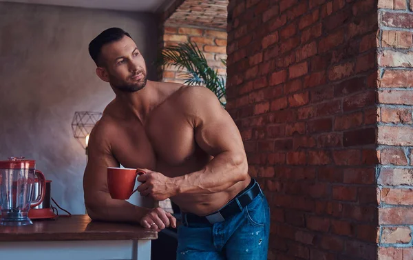 Portrait of a Tall stylish shirtless bodybuilder dressed in jeans, enjoys the morning invigorating coffee, standing in a kitchen with a loft interior, looking out the window.