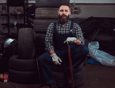 Garajda eski araba lastikleri tutan bir levye, üniforma giymiş sakallı makinist oturur.