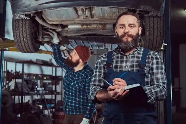 Bearded expert mechanic records results of inspecting a car while his partner checking a problem with a car suspension. A scene from a service station.