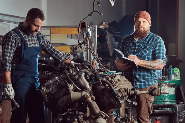 Bearded expert mechanic records results of inspecting a car while his partner checking a problem with a car engine. A scene from a service station.