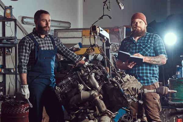 Bearded expert mechanic records results of inspecting a car while his partner checking a problem with a car engine. A scene from a service station.