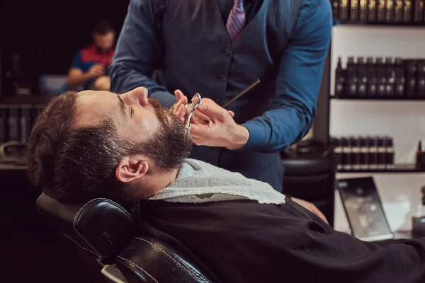 Bearded male sitting in an armchair in a barber shop while hairdresser modeling beard with scissors and comb at the barbershop.