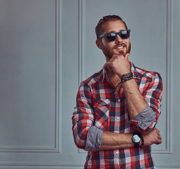 A handsome smiling stylish redhead man in a flannel shirt and sunglasses, posing in a studio against a white wall.