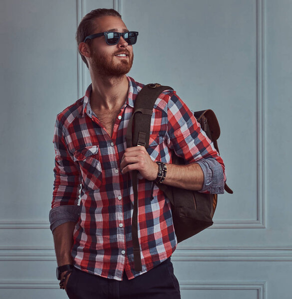 Красивый улыбающийся стильный рыжий путешественник в фланелевой рубашке и солнцезащитных очках с рюкзаком, позирующий в студии у белой стены
.