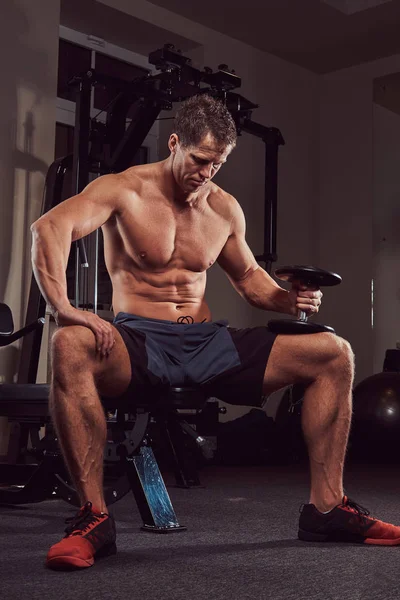 一个肌肉不穿上衣的运动员坐在健身房的长凳上做哑铃运动 — 图库照片