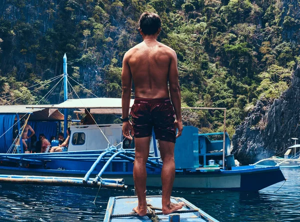 Shirtless Asian guy standing on a bow of the ship on the background of a beautiful landscape of rocky bay.
