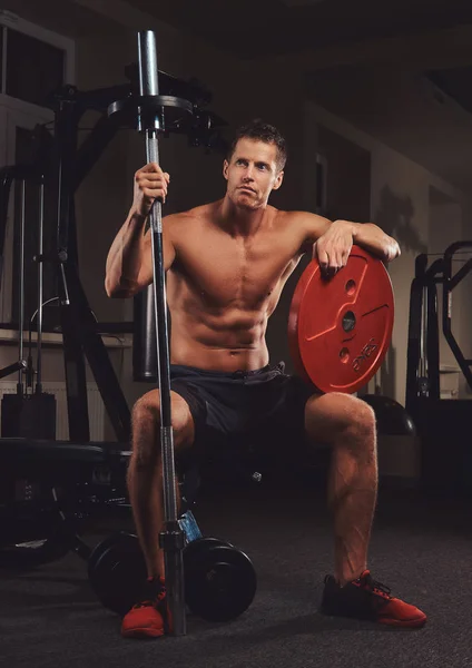 Gespierde shirtless atleet houdt barbell schijf terwijl zit op een bankje in de sportschool. — Stockfoto