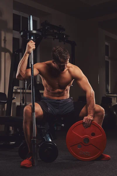 Atleta musculoso sin camisa sostiene el disco de la barra mientras se sienta en un banco en el gimnasio . — Foto de Stock