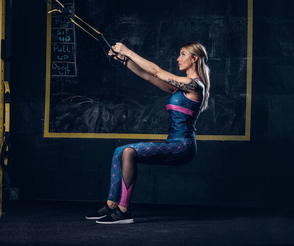 Beautiful muscular blonde woman in sportswear with a tattoo on her arm doing exercise with TRX system at gym. TRX concept.