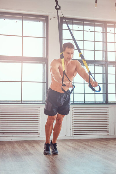 Shirtless muscular male doing exercise with TRX. Functional workout at home with TRX loops.