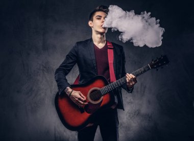 Yakışıklı genç müzisyen şık giysileriyle şık saç ile akustik gitar çalarken duman exhales. Karanlık bir arka plan üzerinde izole.