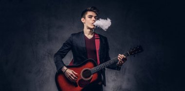 Yakışıklı genç müzisyen şık giysileriyle şık saç ile akustik gitar çalarken duman exhales. Karanlık bir arka plan üzerinde izole.