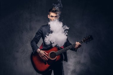 Yakışıklı genç müzisyen şık giysileriyle şık saç ile akustik gitar çalarken duman exhales. Karanlık bir arka plan üzerinde izole.