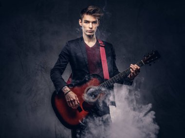 Yakışıklı genç müzisyen şık giysileriyle şık saç ile akustik gitar çalarken duman exhales. Karanlık bir arka plan üzerinde izole.