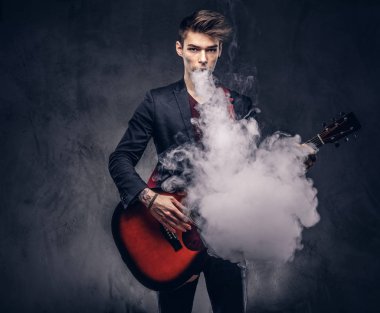 Yakışıklı genç müzisyen şık giysileriyle şık saç ile akustik gitar çalarken duman exhales. Karanlık bir arka plan üzerinde izole.