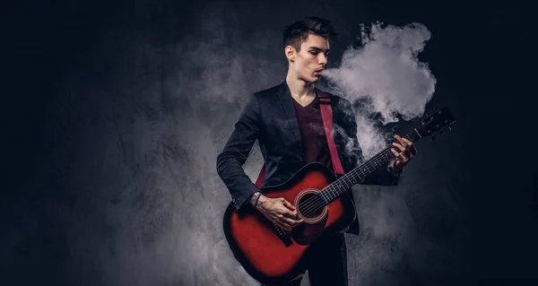 エレガントな服のスタイリッシュな髪のハンサムな若いミュージシャンがアコースティック ギターを演奏しながら煙を吐き出します 暗い背景に分離 — ストック写真