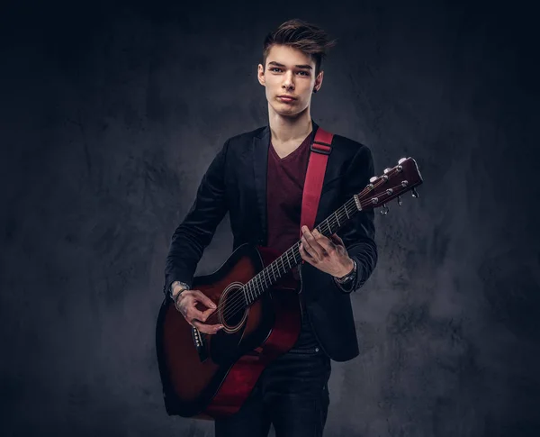 英俊的年轻音乐家与时尚的头发在优雅的衣服与吉他在他的手演奏和摆在黑暗的背景下 — 图库照片