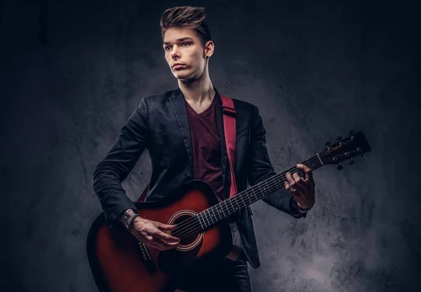 时尚的年轻音乐家与时尚的头发在优雅的衣服与吉他在他的手演奏和摆在黑暗的背景下 — 图库照片