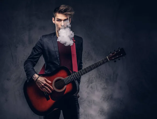 Handsome young musician with stylish hair in elegant clothes exhales smoke while playing acoustic guitar. Isolated on a dark background.
