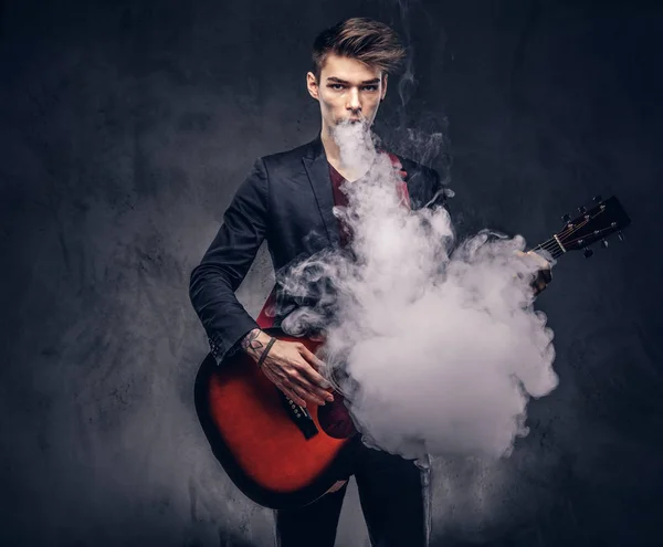 エレガントな服のスタイリッシュな髪のハンサムな若いミュージシャンがアコースティック ギターを演奏しながら煙を吐き出します 暗い背景に分離 — ストック写真