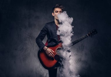 Şık giysileriyle exhales akustik gitar çalarken duman içinde şık saçlı yakışıklı genç müzisyen.