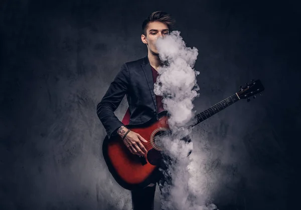 Knappe jonge muzikant met stijlvolle haren in elegante kleding uitademt rook tijdens het spelen van akoestische gitaar. — Stockfoto