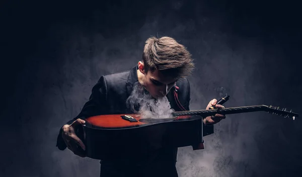 Knappe jonge muzikant met stijlvolle haren in elegante kleding uitademt rook op zijn akoestische gitaar. — Stockfoto