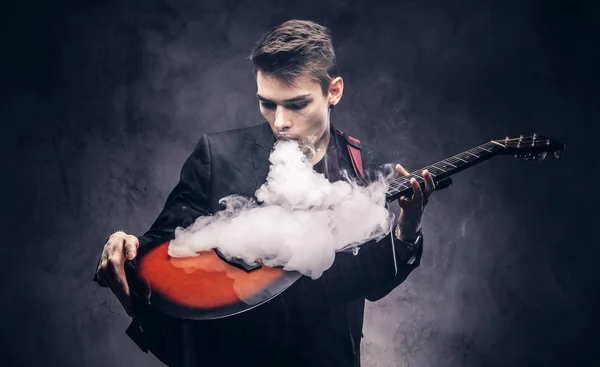 Knappe jonge muzikant met stijlvolle haren in elegante kleding uitademt rook op zijn akoestische gitaar. — Stockfoto
