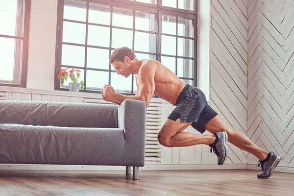 Hombre musculoso guapo sin camisa hace ejercicio apoyado en un sofá en casa . — Foto de Stock