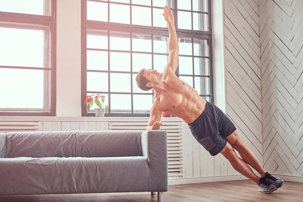 Handsome shirtless muscular male does exercise leaning on a sofa at home. — Stock Photo, Image