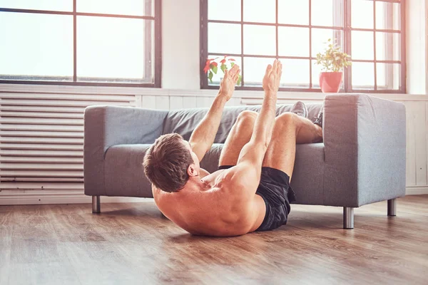 Hombre musculoso guapo sin camisa haciendo ejercicios abdominales en el suelo en casa., apoyado en un sofá . — Foto de Stock