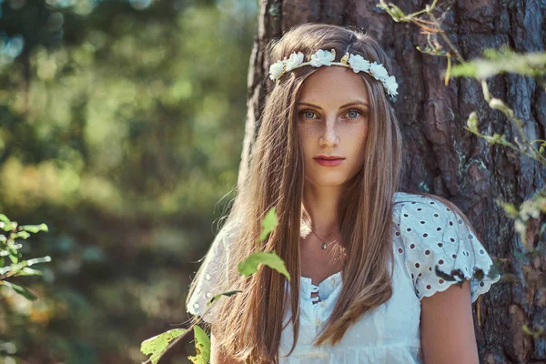 En vacker kvinna i en vit klänning och vit krans på huvudet poserar i en grön höst skog. — Stockfoto