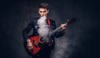 Şık giysileriyle exhales akustik gitar çalarken duman içinde şık saçlı yakışıklı genç müzisyen.