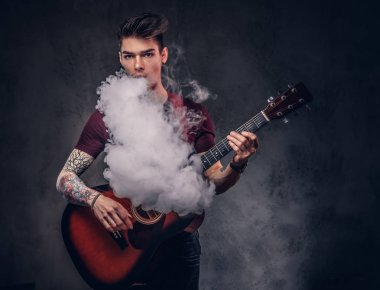 Bir t-shirt, şık saçlı yakışıklı genç müzisyen exhales duman akustik gitar çalarken.